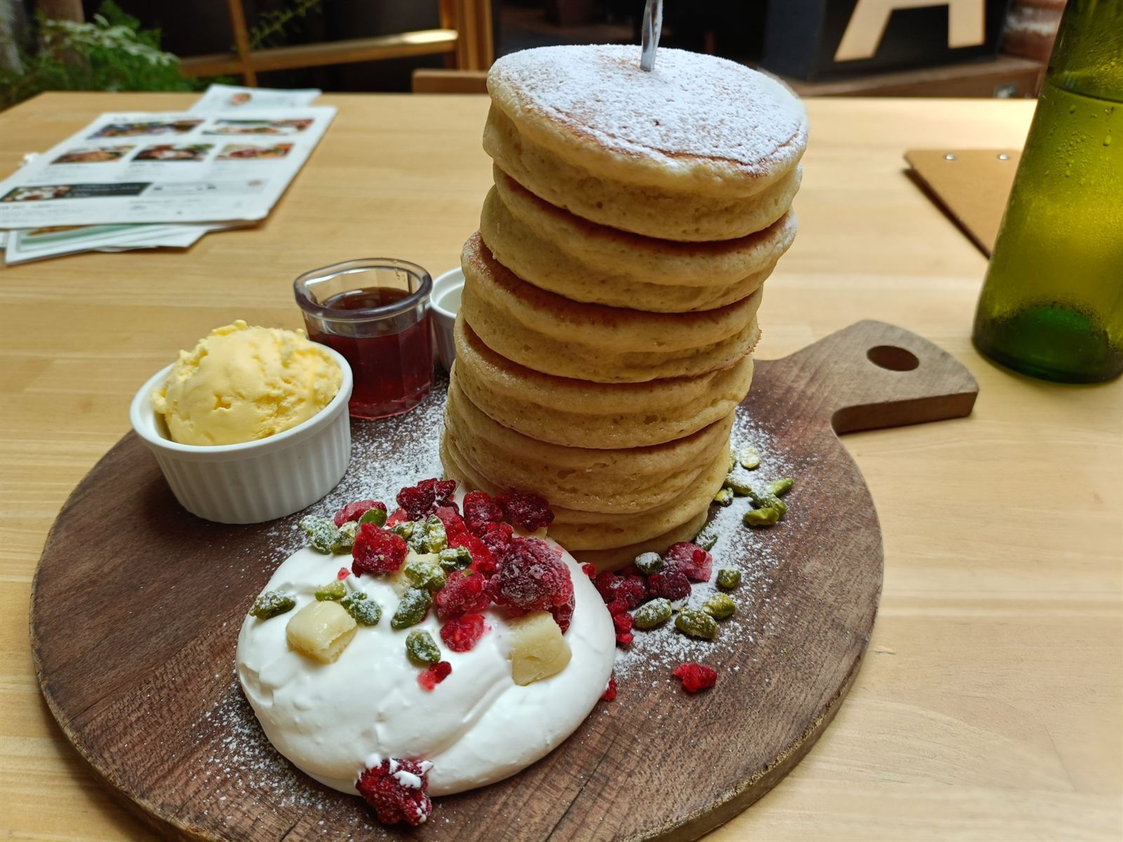 木のボードの上に高く積み重なったパンケーキ。ホイップクリーム、ベリー、ピスタチオ、アイスとシロップが添えられている